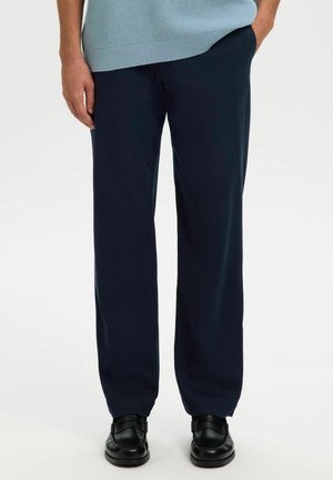 Homme portant un pantalon bleu marine foncé à jambes droites, des chaussures en cuir noires et un pull en maille bleu clair, les mains dans les poches.