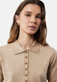 Beige stickad polo med korta ärmar, en mjuk krage och en knappslå med runda, ljusbruna knappar.