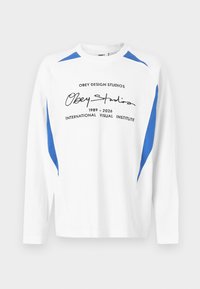 Άσπρο μακρυμάνικο μπλουζάκι με στρογγυλή λαιμόκοψη, με μπλε λεπτομέρειες στους ώμους και στα πλαϊνά, με μαύρο κείμενο "Obey Studios 1989-2026 International Visual Institute" μπροστά.