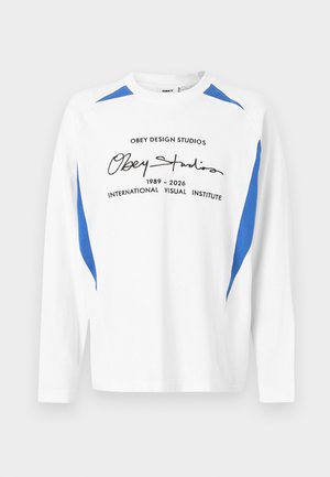 Weißes Langarm-Shirt mit Rundhalsausschnitt und blauen Akzenten an Schultern und Seiten, mit schwarzem Text "Obey Studios 1989-2026 International Visual Institute" auf der Vorderseite.