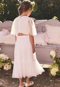 Vestido blanco con falda de tul en capas, mangas cortas de capa transparente y cinturilla elástica. Zapatos dorados. El fondo incluye flores blancas y un banco.