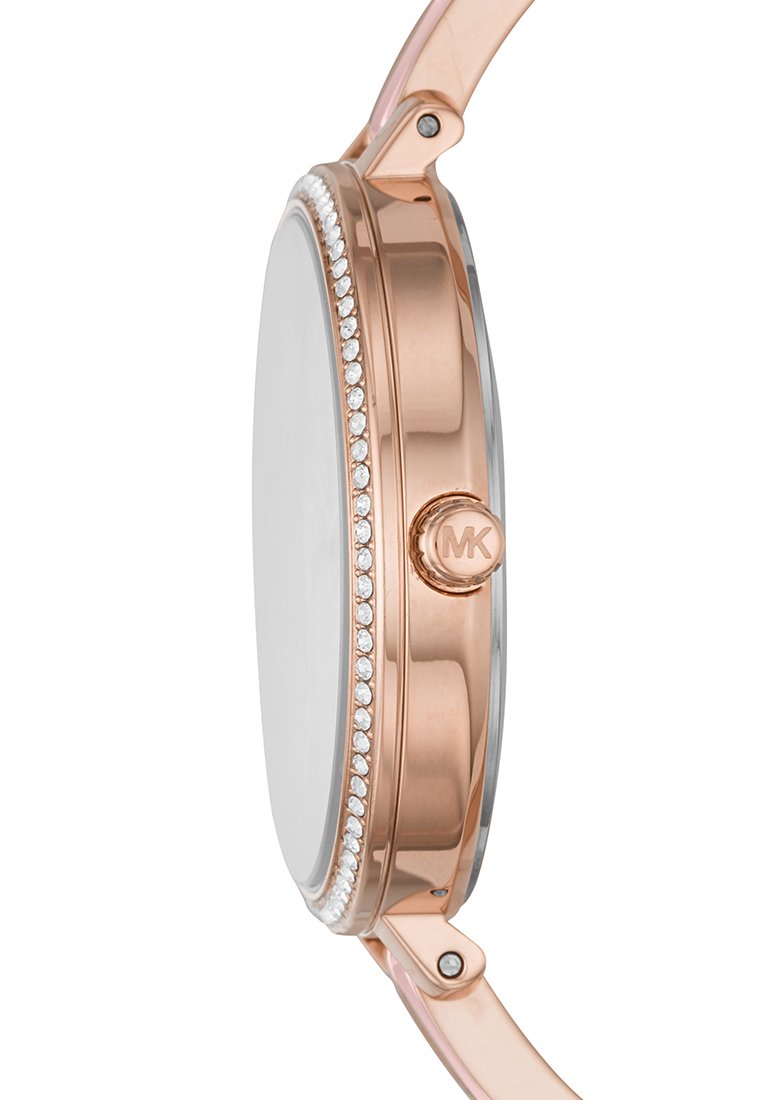 Reloj de oro rosa visto de lado con bisel adornado con cristales y corona con el logo de MK, unido a una correa de oro rosa a juego.