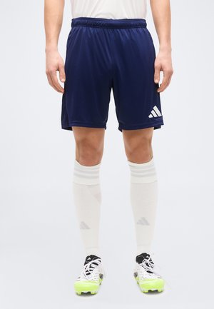 Άνδρας που φοράει σορτς Adidas σκούρου μπλε, λευκές κάλτσες μέχρι το γόνατο και λευκά παπούτσια ποδοσφαίρου με φωσφορίζουσες κίτρινες σόλες, στέκεται μπροστά σε ένα απλό φόντο.