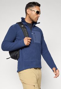 Giacca in pile blu navy con tessuto testurizzato, tasca sul petto con zip, colletto alto e maniche lunghe; indossata con uno zaino nero.