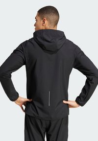 Schwarze Performance-Jacke mit Kapuze, die ein elegantes Design, lange Ärmel und ein reflektierendes Reißverschlussdetail auf dem Rücken aufweist.