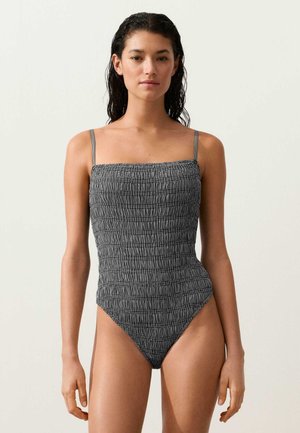 Žena s mokrými vlasmi, oblečená v čierno-bielych štruktúrovaných jednočastných plavkách s tenkými ramienkami, tvárou vpred na jednofarebnom pozadí.