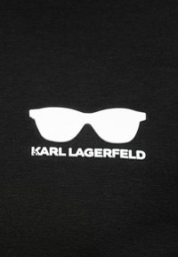 Λευκό περίγραμμα γυαλιών ηλίου πάνω από το κείμενο "KARL LAGERFELD" σε μαύρο ύφασμα, που αντιπροσωπεύει το λογότυπο μιας μόδας μάρκας.
