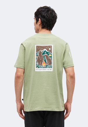 Uomo che indossa una maglietta verde chiaro con grafica di due orsi vicino al fuoco da campo, montagne, alberi, stelle e la scritta "Vai a dormire sotto il cielo" sulla schiena.