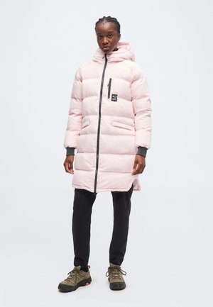 Persona che indossa un lungo piumino rosa chiaro con cappuccio, pantaloni neri e scarpe da trekking marroni, in piedi davanti a uno sfondo bianco uniforme.