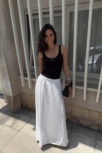 Un débardeur noir et une jupe maxi en lin blanche, avec une taille froncée. Tenue d'une petite pochette tissée noire. Se tenant contre un mur de couleur claire.
