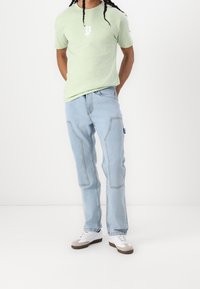 Light green short-sleeve t-shirt with a white graphic print, paired with light blue jeans featuring patch pockets and a straight fit. White and tan sneakers.