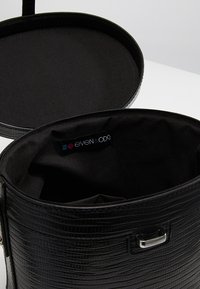 Musta sylinterimäinen käsilaukku, jossa on teksturoidun krokotiilinnahka-efekti. Sisältää sileän, satiinivuorauksen. Varustettu hopeanvärisellä sulkimella.