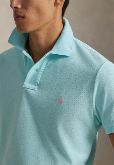 Ανοιχτό μπλε πόλο μπλουζάκι σε υφασμάτινη υφή, με ροζ κεντητό λογότυπο στο αριστερό στήθος και κλασικό γιακά με δύο κουμπιά.