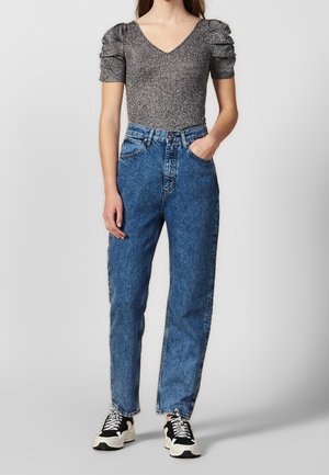 Femme portant un haut argenté à manches bouffantes, un jean bleu taille haute avec une coupe décontractée et des baskets noires et blanches, debout avec une main dans la poche.