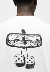 Πίσω όψη ατόμου που φοράει λευκό πουκάμισο με μαύρο γραφικό που απεικονίζει τον καθρέφτη οπισθοπορείας αυτοκινήτου, που δείχνει έναν καουμπόι επάνω σε άλογο στην έρημο και κρεμαστά μαλακά ζάρια.