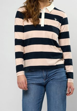 Femme portant un polo à manches longues avec de larges rayures horizontales bleu marine et rose clair, associé à un jean bleu, debout devant un fond uni.