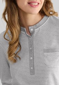 Close-up de una persona que lleva una camisa de rayas horizontales grises y blancas con botones y un pequeño bolsillo en el pecho, sonriendo levemente.