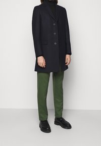 Manteau navy avec un design structuré, fermeture simple boutonnée et trois boutons. Associé à un pantalon droit vert et des chaussures noires.