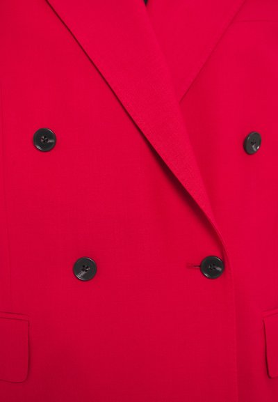 Blazer rouge à double boutonnage avec une texture lisse, présentant des boutons noirs et des poches latérales, mettant en avant un design structuré.