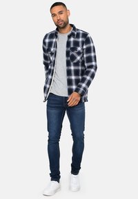 Marineblauw en witte geruite flanellen shirt met twee voorzakken, gedragen over een lichtgrijs T-shirt, gecombineerd met donkere spijkerbroeken en witte sneakers.