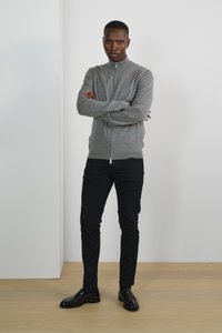 Grå zip-up tröja med hög krage, kombinerad med svarta slim-fit byxor och glänsande svarta skor, stående på ett trägolv mot en ljus vägg.