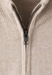 Beige stickad tröja med hög krage, utrustad med en svart dragkedja och ribbat detaljer längs halsringningen. Tvärrad, mjukt material.