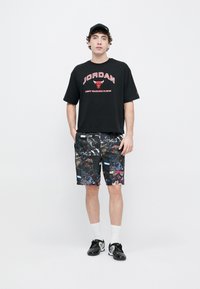 Μαύρο βαμβακερό T-shirt με το λογότυπο "JORDAN" και κόκκινες λεπτομέρειες· μαύρο σορτς με πολύχρωμα γραφικά μπάσκετ· μαύρα παπούτσια και λευκές κάλτσες.