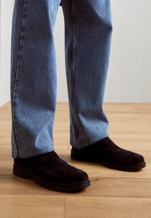 Nogi w jasnoniebieskich dżinsach z denimu i czarne zamszowe buty wsuwane stojące na drewnianej podłodze na jednolitym tle.