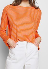 Orange och grå randig långärmad skjorta med avslappnad passform, i kombination med vita högmidjade byxor med framfickor.