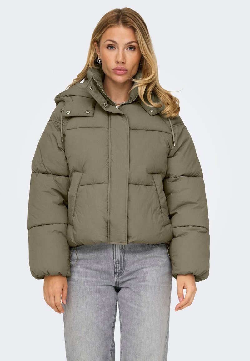 Donna bionda che indossa un giubbotto imbottito verde oliva con cappuccio e jeans grigi chiari, in piedi su uno sfondo chiaro e uniforme.