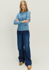 Donna in piedi con le braccia incrociate, che indossa un maglione azzurro chiaro a lavorazione a trecce, jeans ampi scuri e ballerine beige, su uno sfondo neutro.