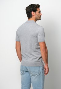 Grå kortärmad pikétröja med stickad textur, krage och ribbat nederkant, som bärs med ljusblå jeans. Betraktarperspektiv från baksidan.