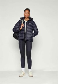 Marineblå puffjakke med glidelås og hette, båret over en grå topp og svarte leggings. Modellen holder en svart ryggsekk, iført hvite sneakers.