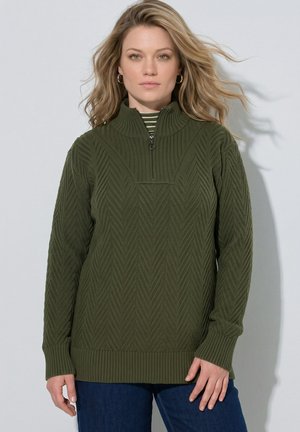 Vrouw met golvend blond haar draagt een groen gestructureerde gebreide trui met halve rits en donkerblauwe jeans, kijkt naar voren tegen een effen achtergrond.