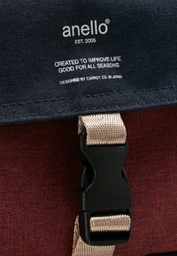 Röd och marinblå tygryggsäck med en svart plastspänne och beige rem, med tryckt varumärke och slogan på locket.