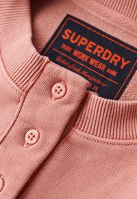Suéter rosa claro con cuello acanalado, que presenta dos botones a juego y una etiqueta azul con "Superdry" en texto naranja.