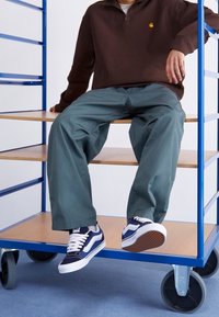 Marineblaue Sneakers mit weißen Akzenten, grüne Hose mit weitem Bein und ein braunes Sweatshirt mit Viertel-Reißverschluss, sitzend auf einem Holzregal.