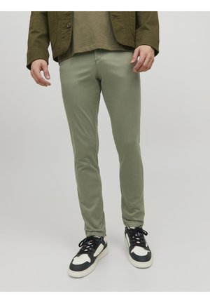 Pantaloni slim-fit verde chiaro realizzati in tessuto morbido, mostrati con una giacca verde scuro e sneakers bianco e nero. Texture liscia e forma su misura.