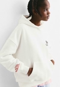 Femme avec des cheveux tressés portant un sweat à capuche blanc surdimensionné avec une poche avant et un texte rouge sur la manche et la poitrine, regardant sur le côté.