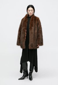 Abrigo de piel sintética marrón con un diseño grueso y texturizado. Llevado sobre un vestido negro con un dobladillo asimétrico y botas negras hasta la rodilla.