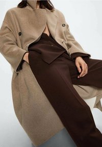 Beige wollen jas met een dubbele rij knopen, voorzien van zwarte knopen en brede revers; gecombineerd met donkerbruine, op maat gemaakte broek.