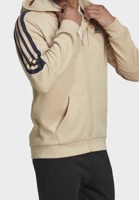 Sweat à capuche beige à fermeture éclair, fabriqué en matériau doux, avec des accents à trois bandes bleu foncé le long des manches et des poches avant.