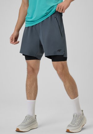Mężczyzna w warstwowych szarych sportowych szortach i białych trampkach stoi na jednolitym jasnoszarym tle.