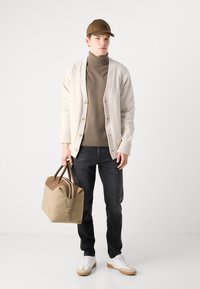 Beige cardigan over brown turtleneck, dark jeans, and white sneakers. Light brown bag with darker accents, standing against a plain background.