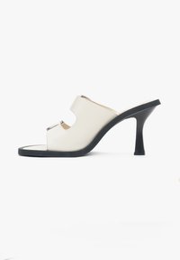 White leather heeled mules with an open toe, featuring curved cutouts and a contrasting black sole. Smooth texture and sleek design.