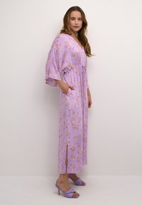Violeta ziedu maxi kleita ar zvana piedurknēm, sašaurinātu vidukli, kabatām un sānu spraugām. Nēsāta ar lavandas krāsas augstpapēžu sandālēm. Gluda tekstūra.
