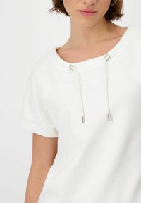 Valkoinen lyhythihainen toppi, jossa on pyöreä pääntie, ja jossa on hopean värisiä yksityiskohtia nyörissä sekä sileä, teksturoitu kangas.