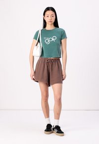 Zelené skrátené tričko s logom "the gap", hnedé textúrované šortky, biela taška, čierne tenisky a biele ponožky. Model stojí na pozadí s jednoduchým vzorom.