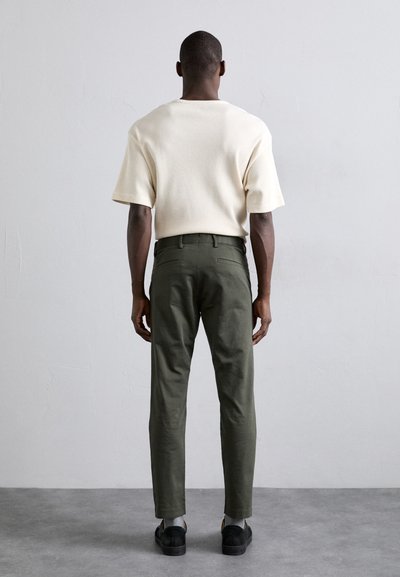 Cremes farbenes, kurzärmliges, strukturiertes Strickshirt kombiniert mit olivgrünen Hosen. Schwarze Schuhe vervollständigen das Outfit. Einfaches, modernes Design.
