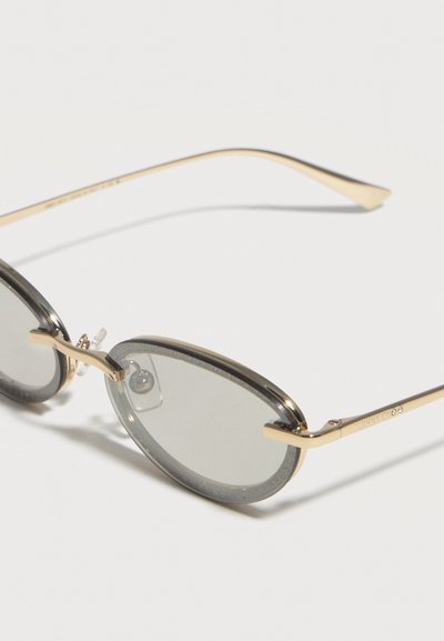 Οβάλ γυαλιά ηλίου με λεπτά χρυσά μεταλλικά πλαίσια και ελαφρώς σκουρόχρωμους γκρι φακούς σε λευκό φόντο.